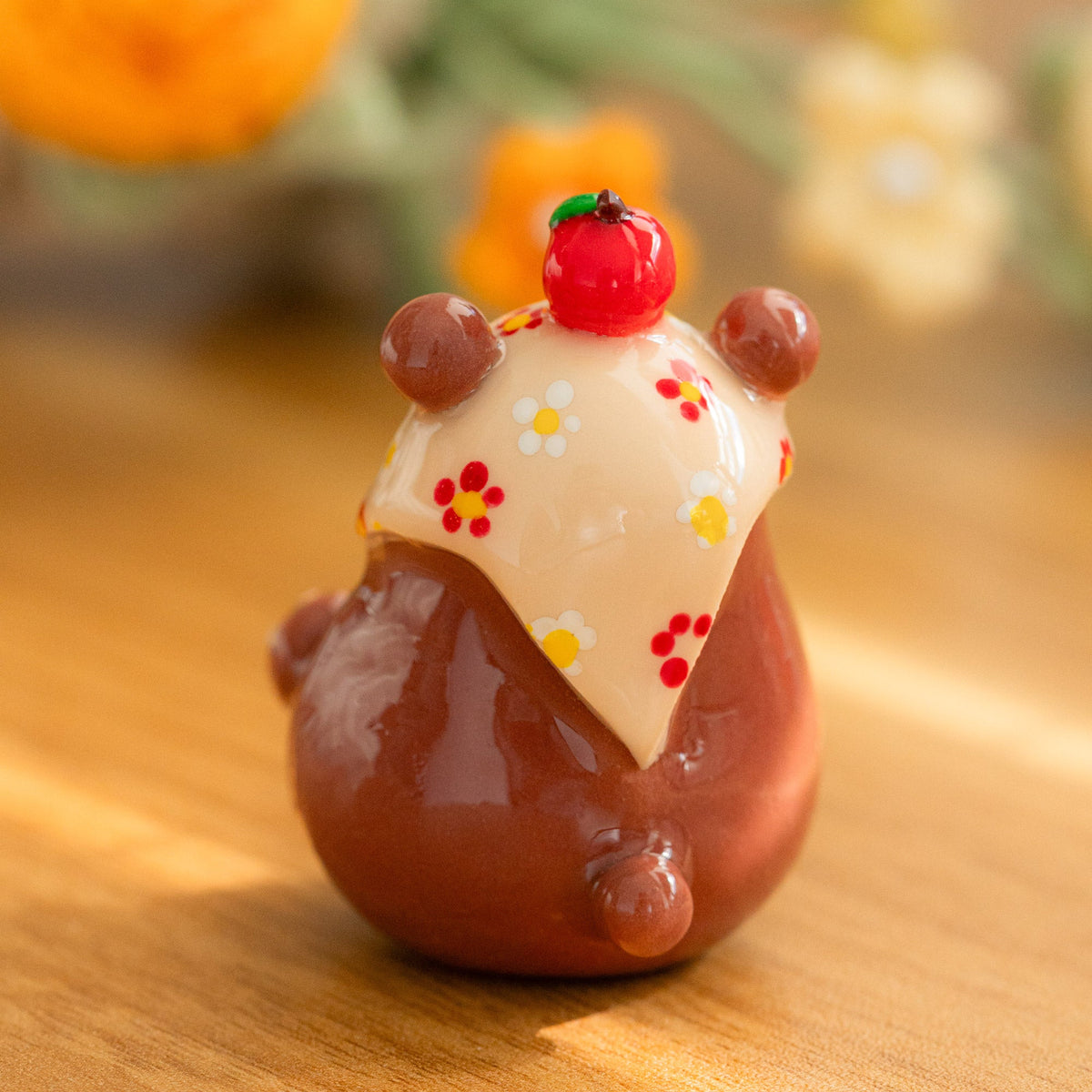 A small glossy brown bear figurine wearing a cream-colored floral scarf and a red apple on top, standing on a warm wooden surface with a softly blurred background.