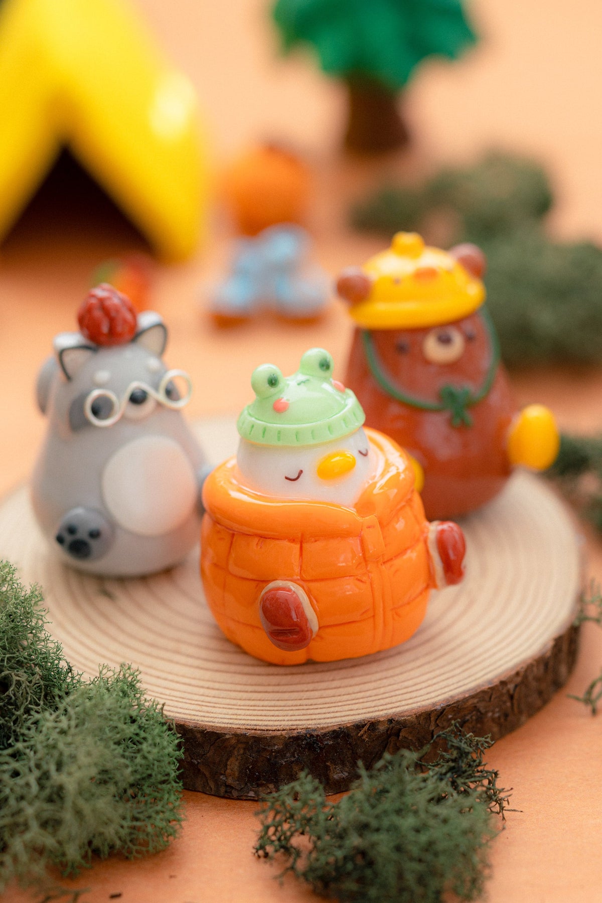 Three cute, kawaii animal figurines displayed on a round wood slice coaster. The central figure is a white duck wearing an orange quilted jacket and a cute green frog hat.