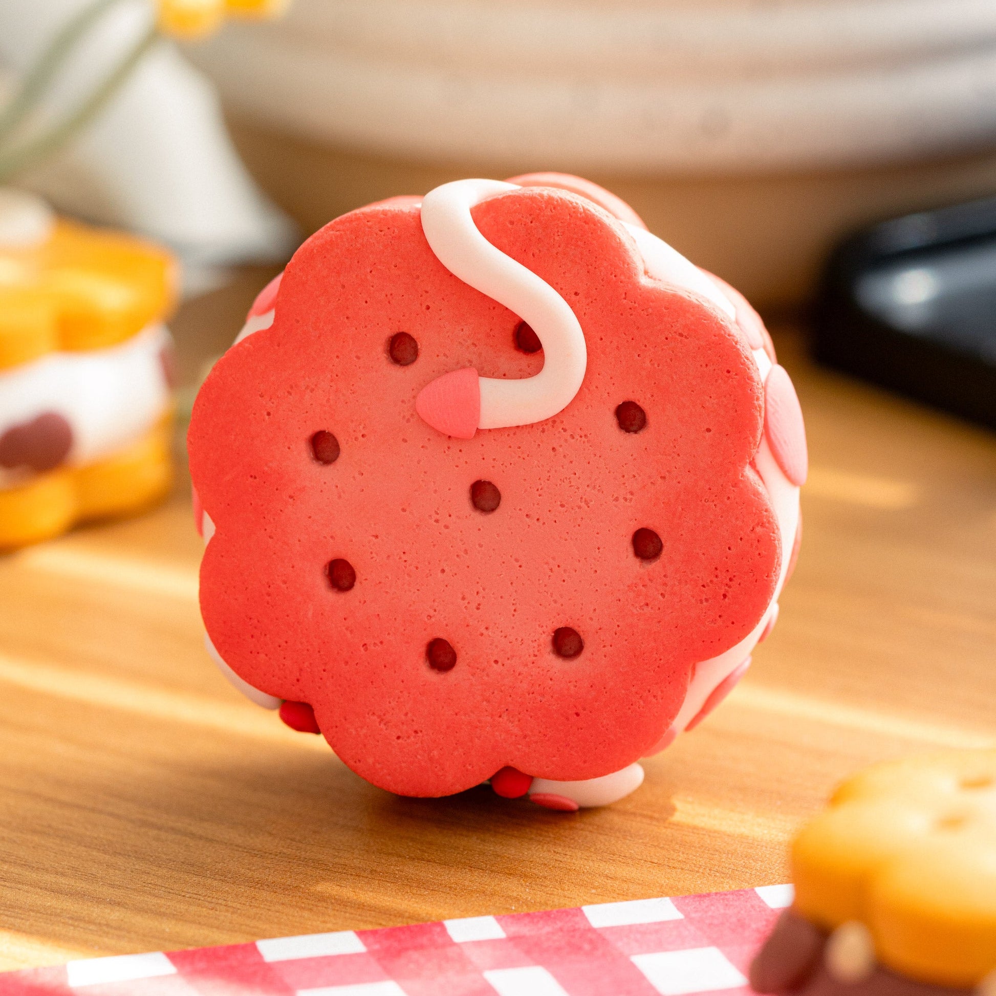 A strawberry-themed cookie-shaped desk buddy, a polymer clay figurine and decorative item, standing upright on a wooden table. The item is strawberry macaron color with white piped details representing strawberry cream and a small pink strawberry accent.