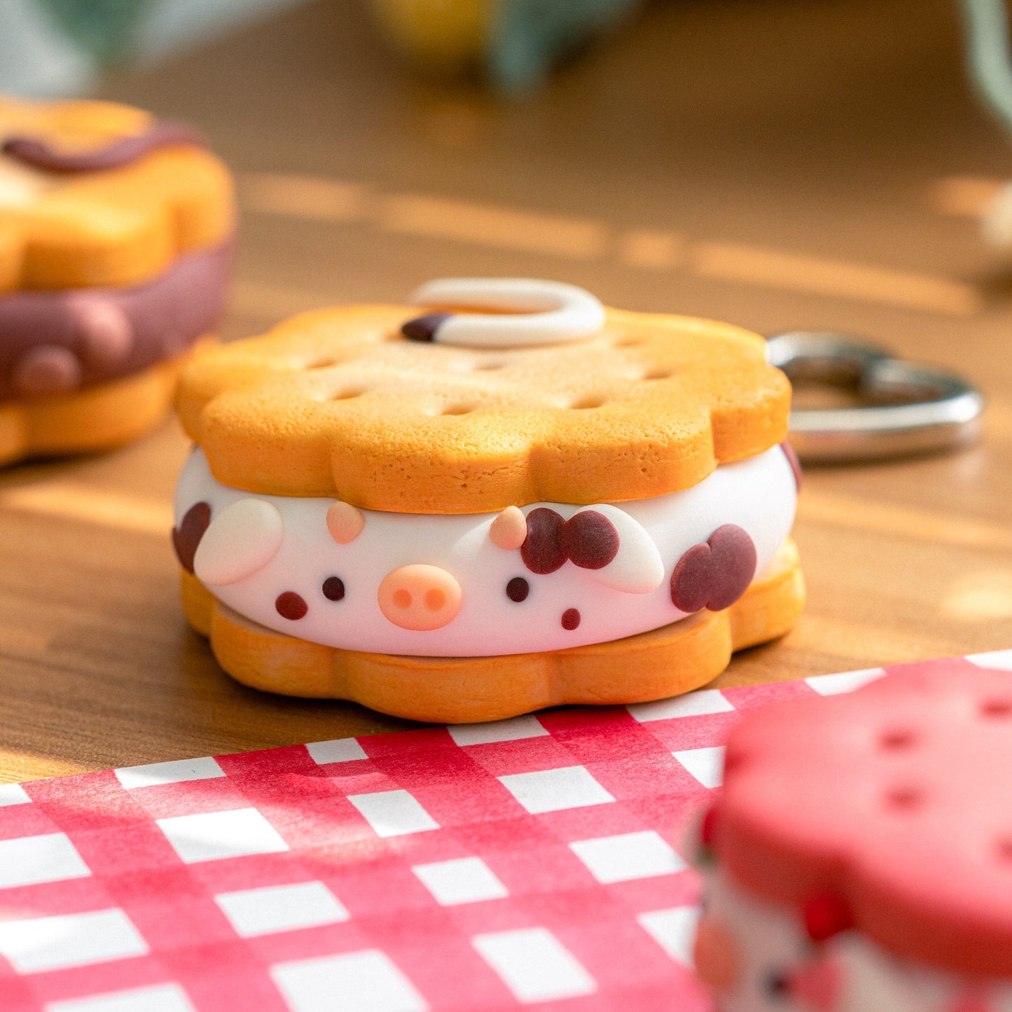 A cute, cow-themed sandwich cookie keychain in the center of the frame, resting on a red and white checkered tablecloth. The cookie features a white cream filling with a printed pig face (snout, ears, and spots) and a silver metal keyring attached.