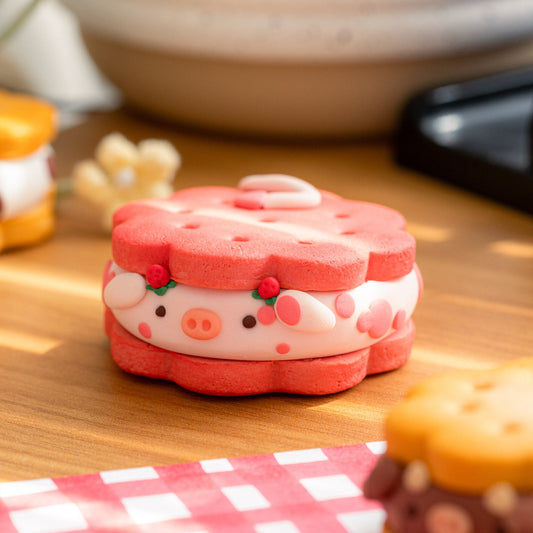 A cute cow-shaped sandwich cookie or clay decoration sits on a wooden table, styled with a red-and-white checkered napkin and other similar treats in the foreground and background.