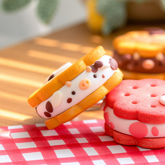 Two cute, cartoon-style sandwich cookies styled like cow faces, one standing upright and the other leaning on a red-and-white checkered napkin on a wooden table.