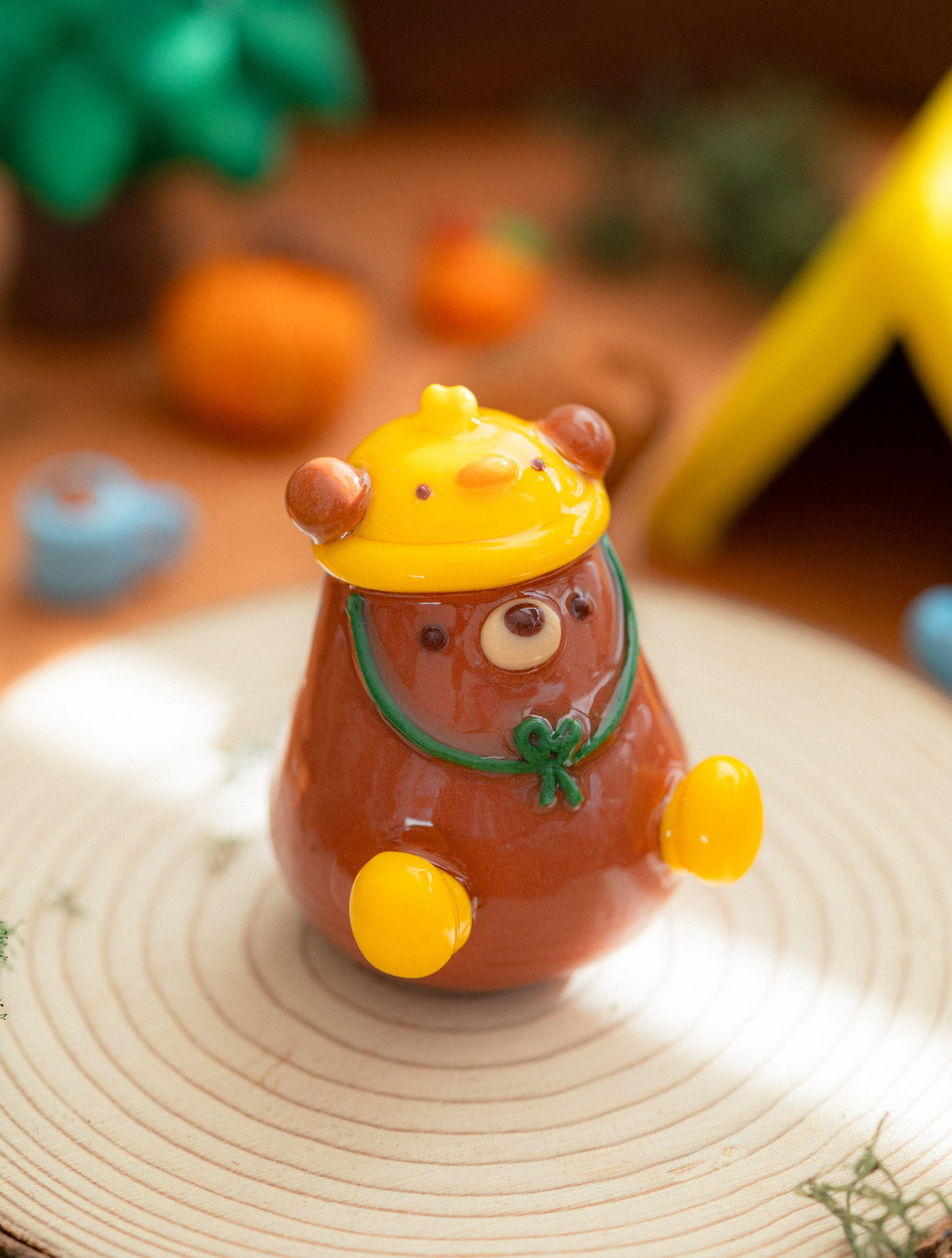 Handmade cozy clay bear figurine with a brown body, yellow Duckie hat and rain boots, displayed on a round wood slice with a blurred autumn-themed background.