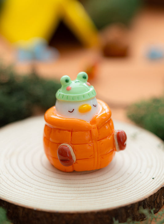 a handmade duck desk figurine sleeping with a kawaii frog hat and a cozy puffer jacket, made from polymer clay. Cozy, kawaii decor.
