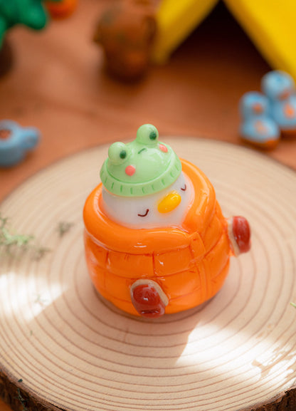 a handmade cozy duck desk figurine with a kawaii frog hat and a warm orange puffer jacket, made from polymer clay. Cozy, kawaii desk decor.