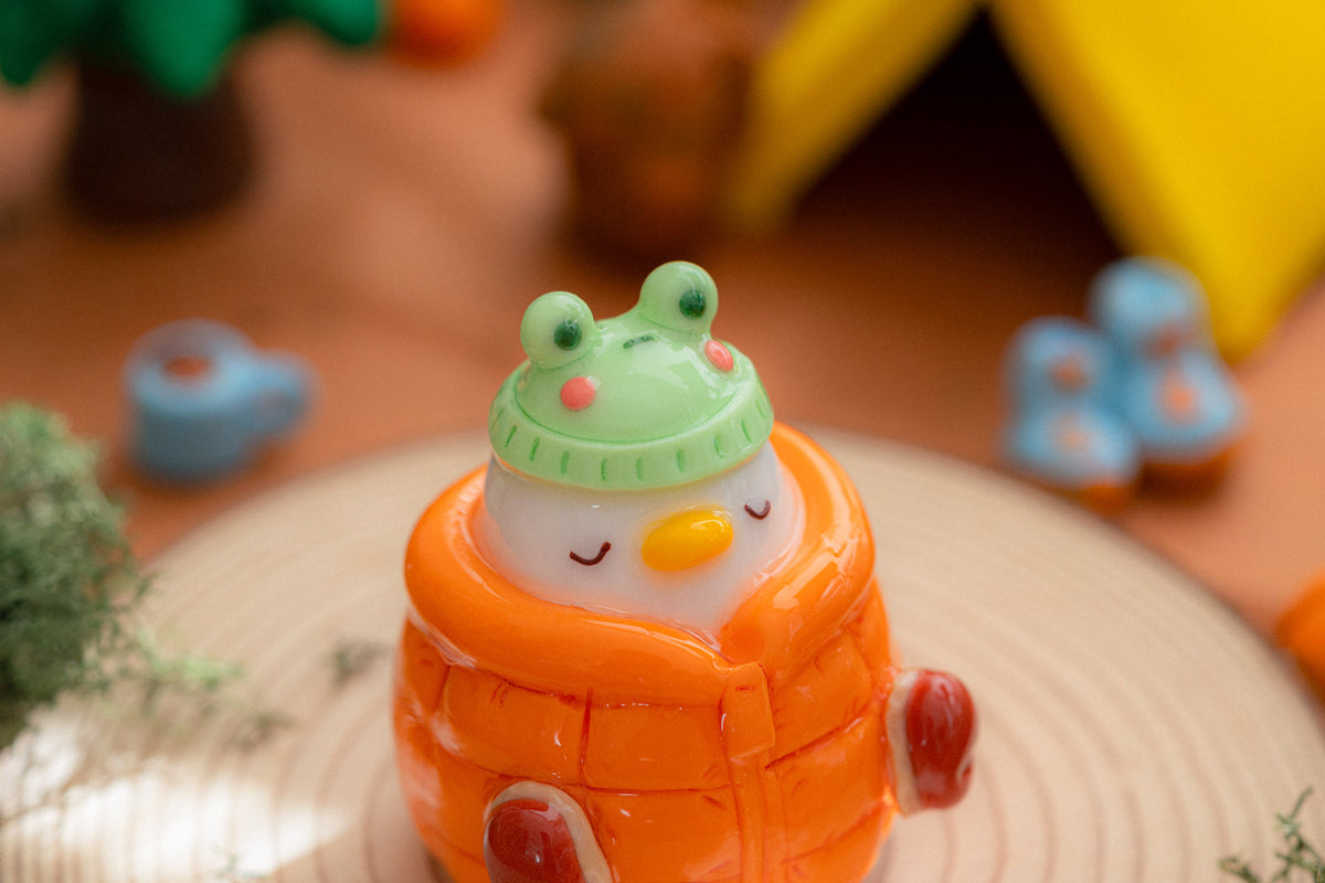 A cute, small decorative figurine shaped like a white duck wearing a bright orange quilted winter coat and a green frog hat, placed on a round wooden plate with a softly blurred cozy camping background.