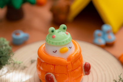 A cute, small decorative figurine shaped like a white duck wearing a bright orange quilted winter coat and a green frog hat, placed on a round wooden plate with a softly blurred cozy camping background.