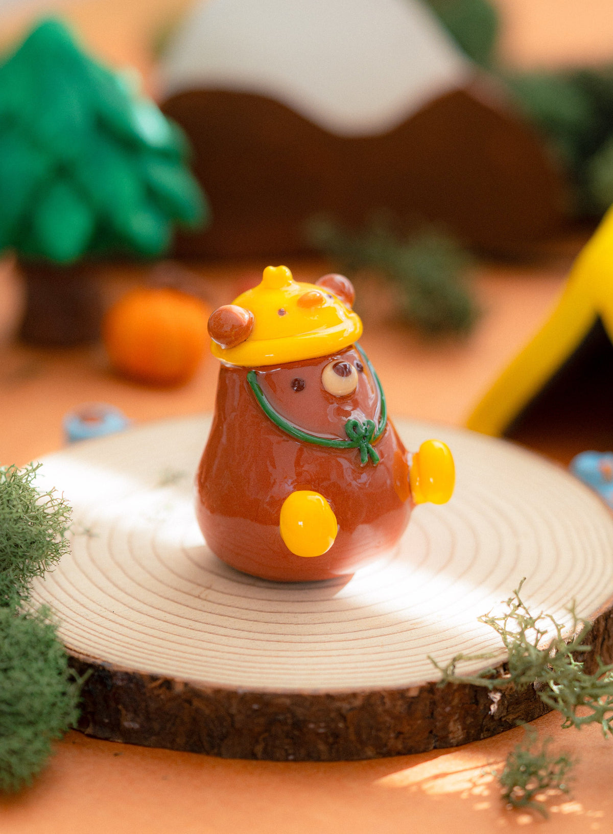 A small clay bear figurine wearing a yellow duck hat with green strings, standing on a round wood log with a softly blurred outdoor-style camping in Colorado scene in the background.