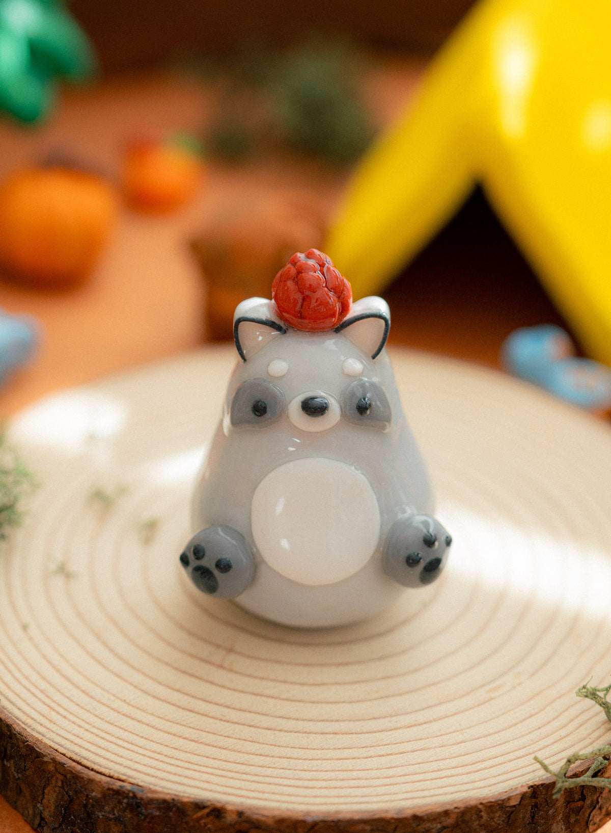 A small gray kawaii-style raccoon figurine with a white belly, dark gray feet and ears, and a brown pinecone on its head, displayed on a round wood slice with a softly blurred warm-toned background.