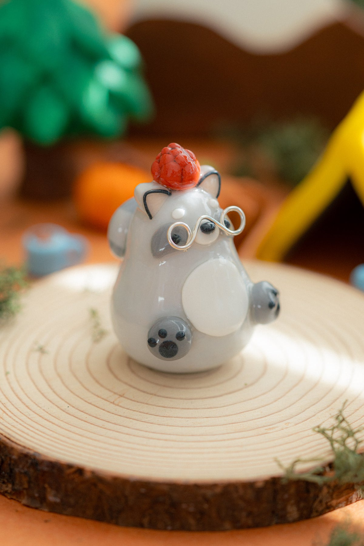 A gray raccoon figurine with a white body, gray ears and paws, round wire glasses, and a pine cone on its head, displayed on a round wood slice with a blurred natural-themed background.