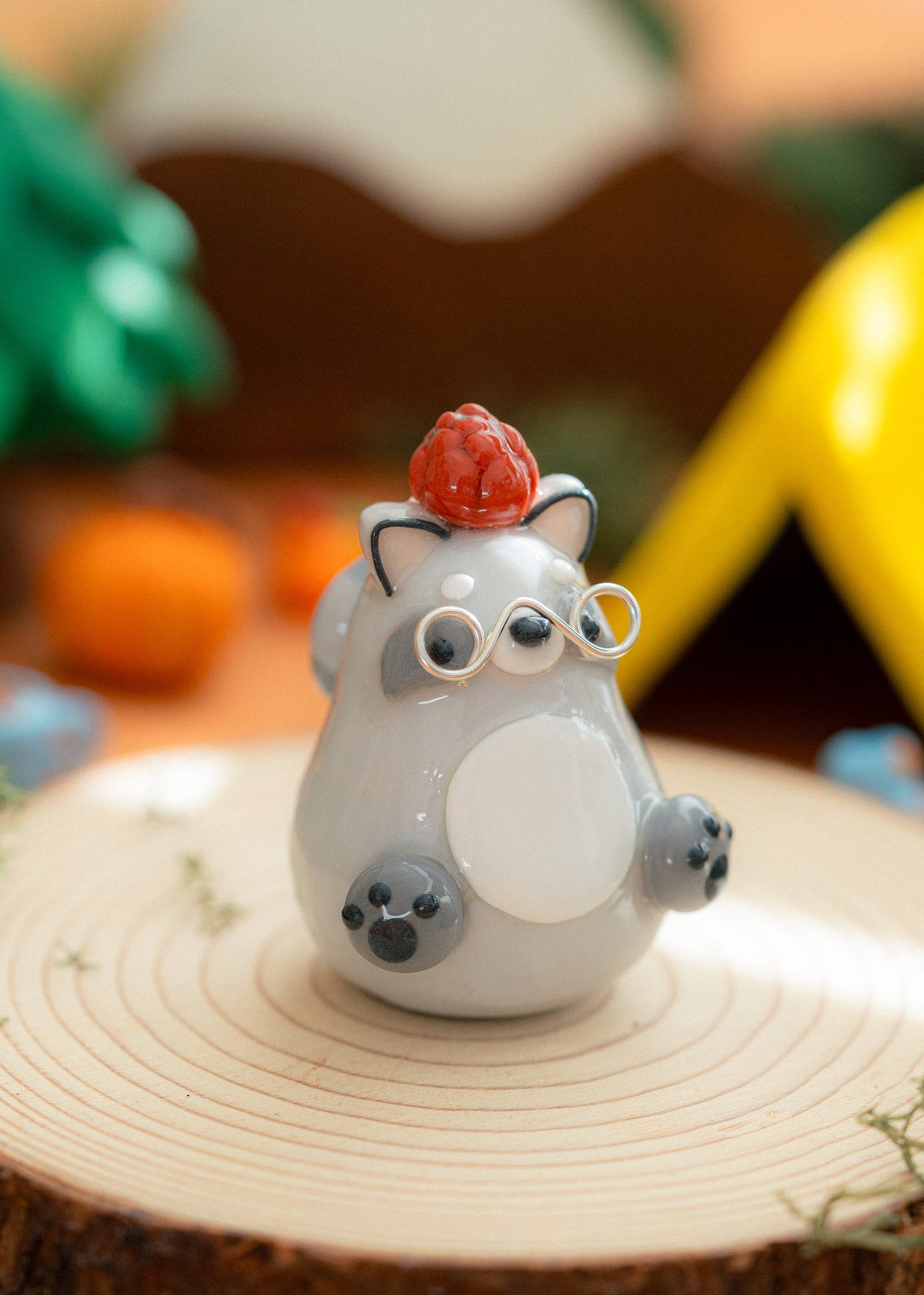 A small kawaii clay raccoon figurine with a brown pine cone on its head, standing on a round wooden slice with silver glasses and a softly blurred outdoor-style background.