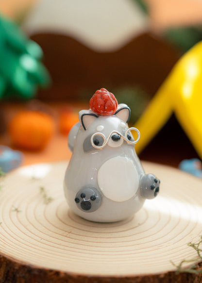 A small kawaii clay raccoon figurine with a brown pine cone on its head, standing on a round wooden slice with silver glasses and a softly blurred outdoor-style background.