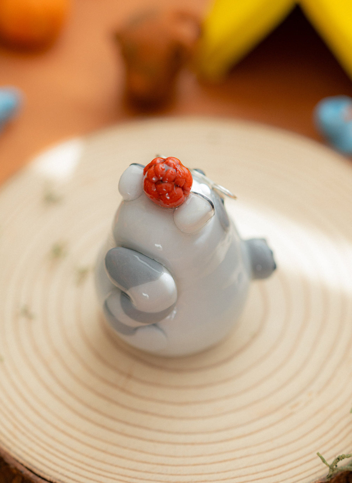 A small glossy clay raccoon figurine holding a pine cone, displayed on a round wooden slice coaster with a warm, softly blurred background.